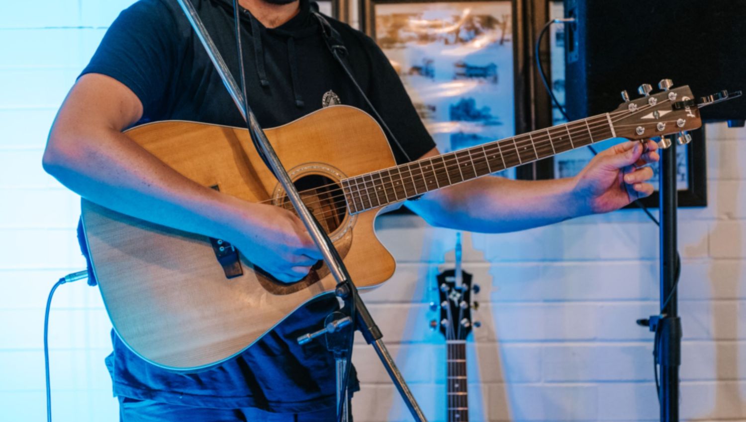 A close-up, cropped shot of a musician holding an acoustic guitar and adjusting the tuning pegs on the headstock during a live performance setup, backlit by blue light.